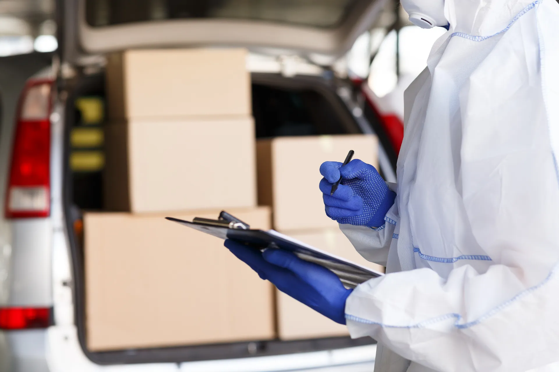 A person in a protective suit and blue gloves holds a clipboard and pen in front of a car packed with cardboard boxes.